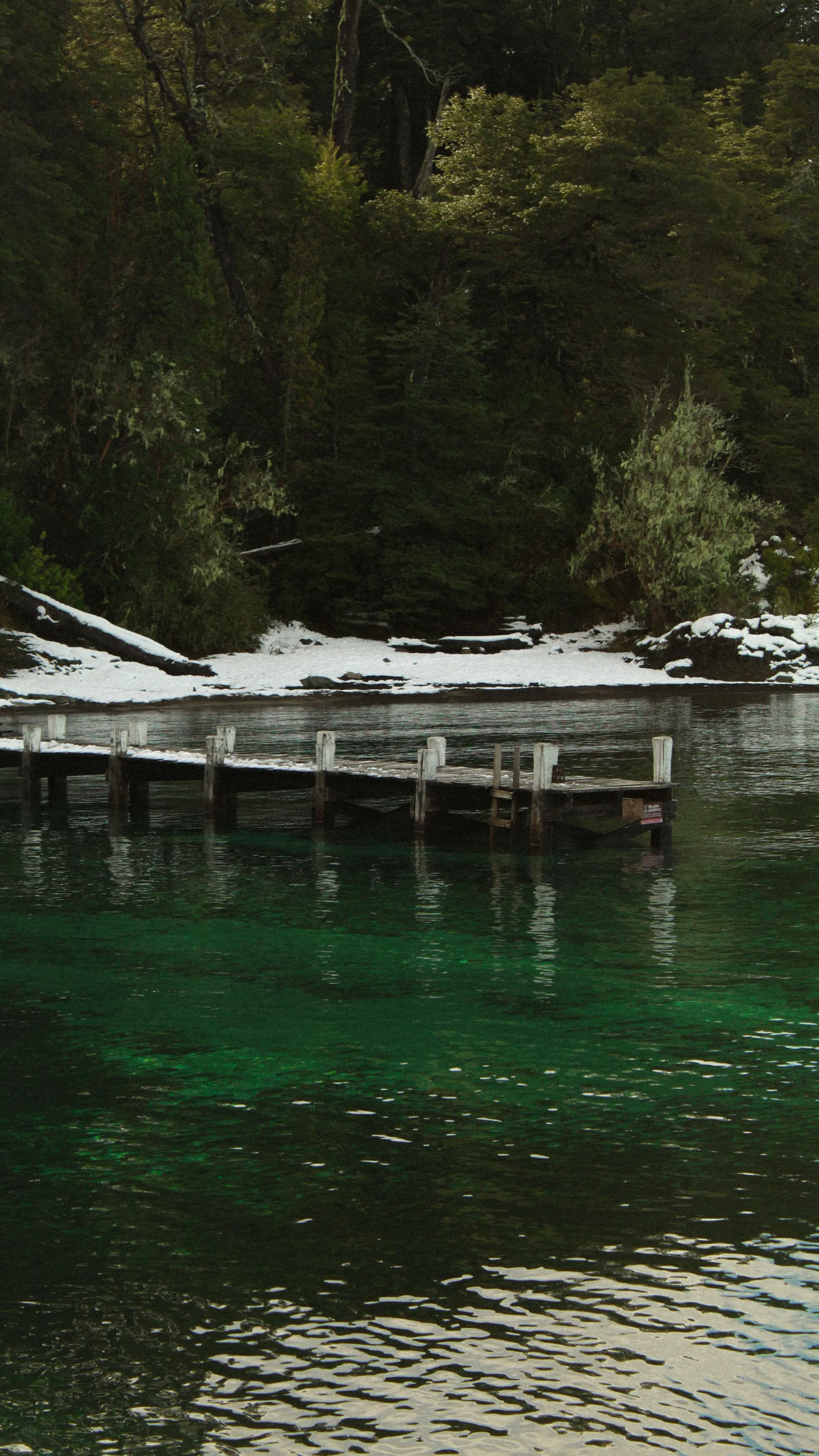 A snow-dusted wooden pier beside green lake water in Villa La Angostura