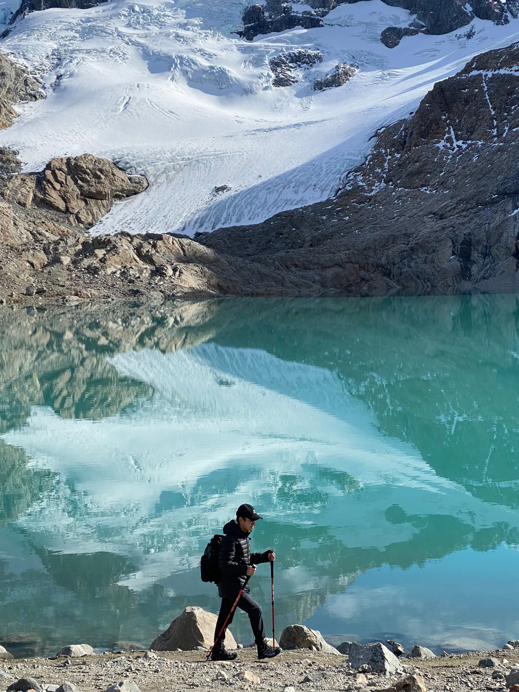 A walker beside a turquoise glacial lake below snowy mountains.